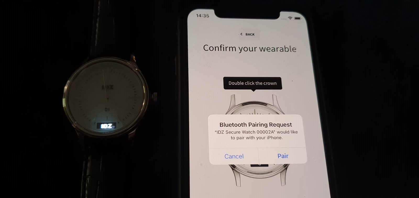 A real IDZ watch prototype beside an iPhone showing the 'Confirm your wearable' pairing screen with a Bluetooth pairing request for 'IDZ Secure Watch'.